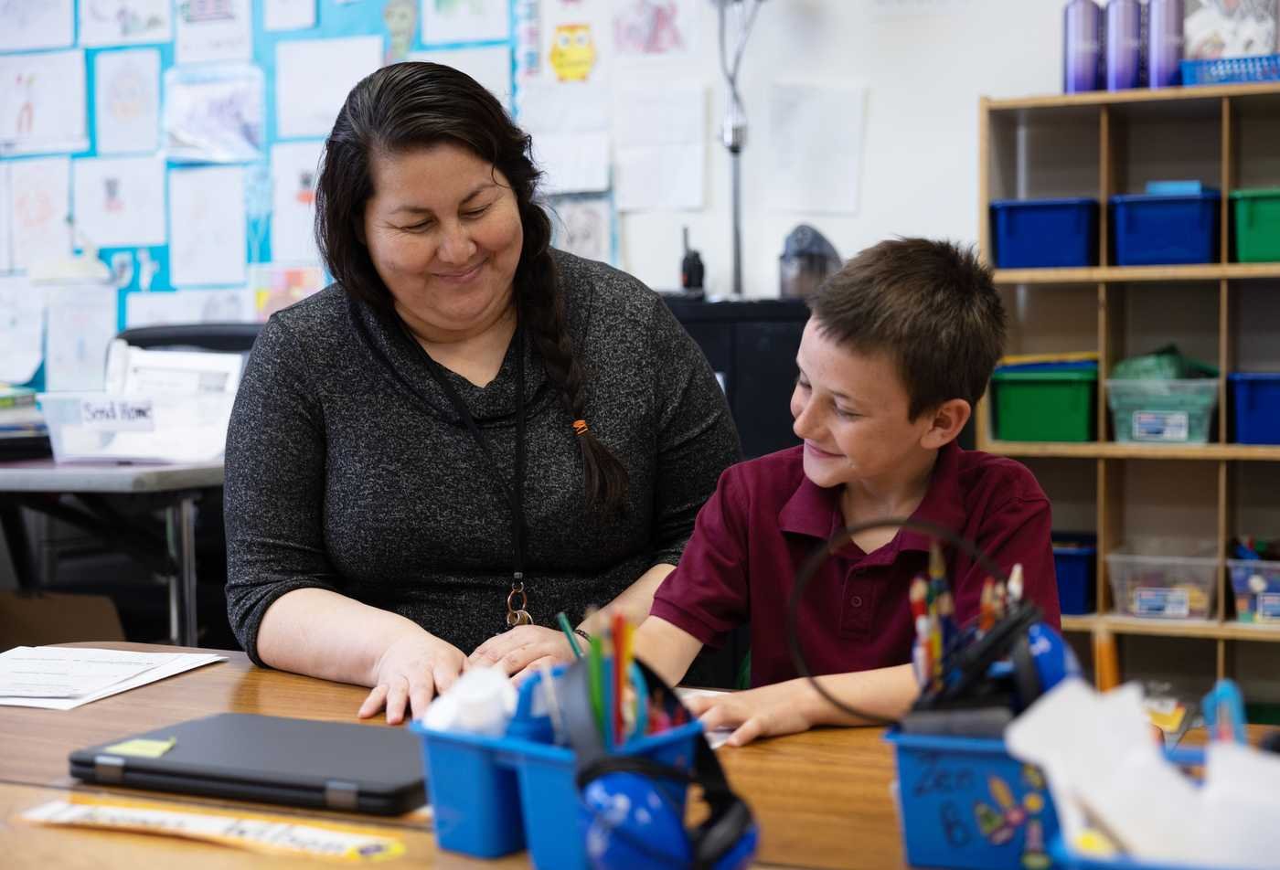 A teacher and a young boy engage in a learning activity at a wooden table in a colorful classroom filled with student artwork, showcasing the expertise gained in the MEd in Special Education (Teacher Certification).