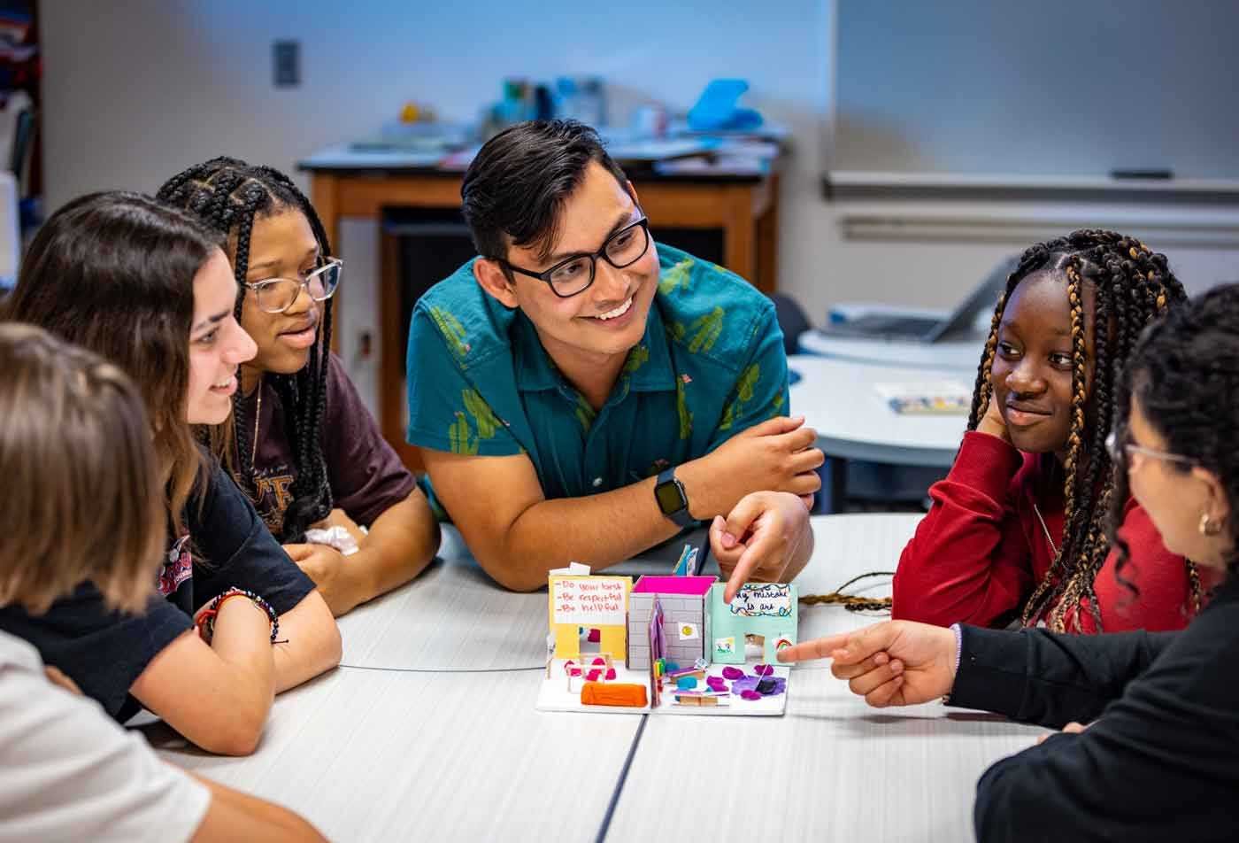 A smiling instructor examines a diarama of a classroom with his students, demonstrating the hands-on learning opportunities in the BAE in Secondary Education.