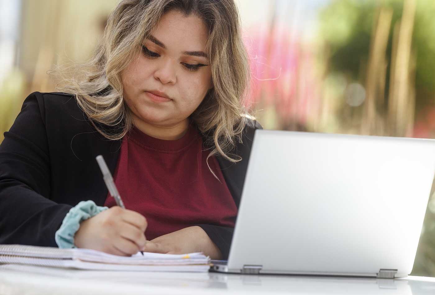 A student writes notes beside a laptop outdoors, developing written communication skills in ASU's Writing certificate.
